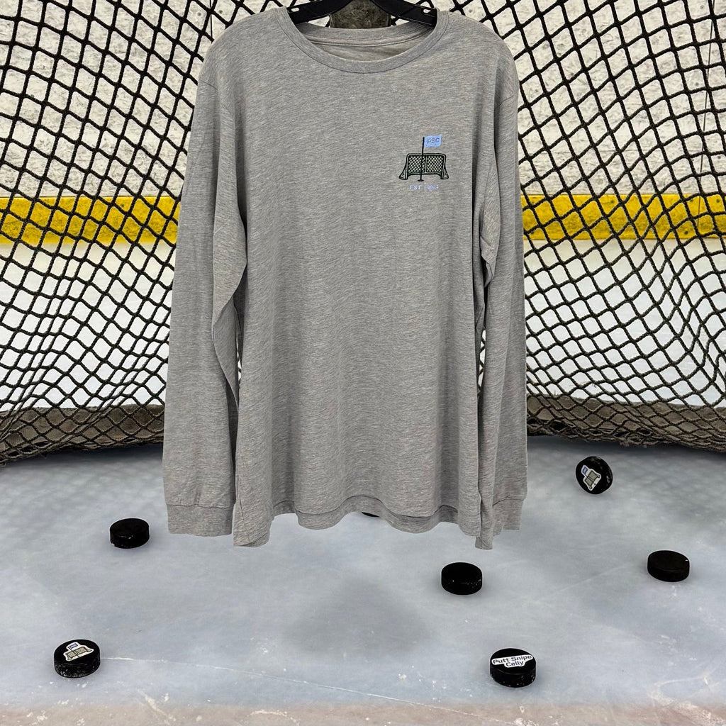 View of the front of the Heritage design in the athletic heather colored long sleeve lifestyle t-shirt shown hanging from a hockey net with pucks on the ice below. Presented by Putt Snipe Celly