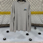 View of the front of the Heritage design in the athletic heather colored long sleeve lifestyle t-shirt shown hanging from a hockey net with pucks on the ice below. Presented by Putt Snipe Celly