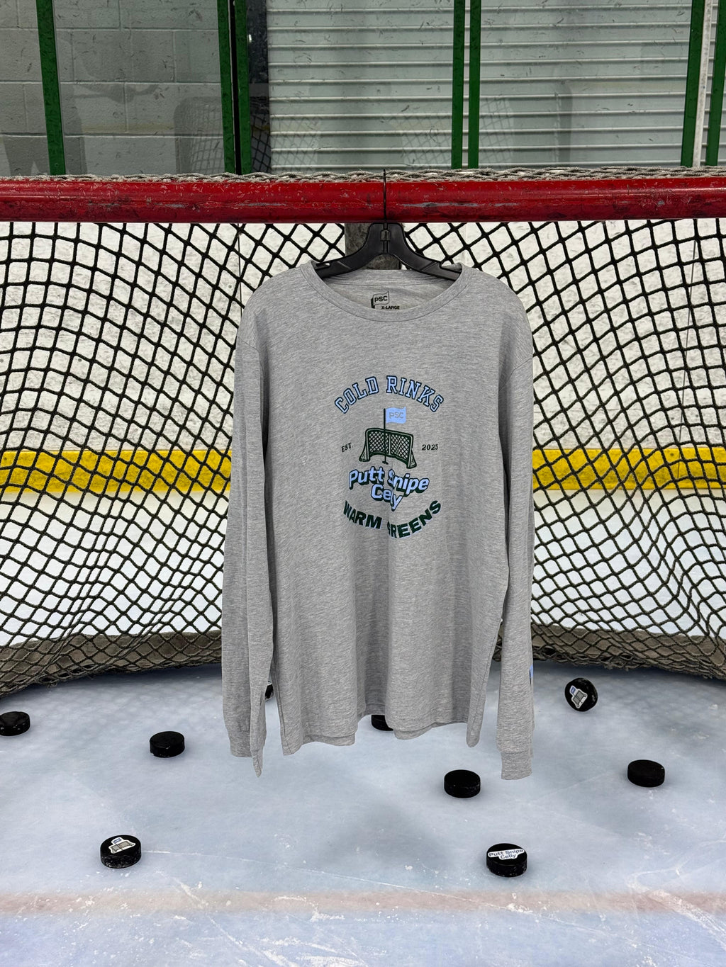Heritage design in the athletic heather colored long sleeve lifestyle t-shirt shown hanging from a hockey net with pucks on the ice below. Presented by Putt Snipe Celly