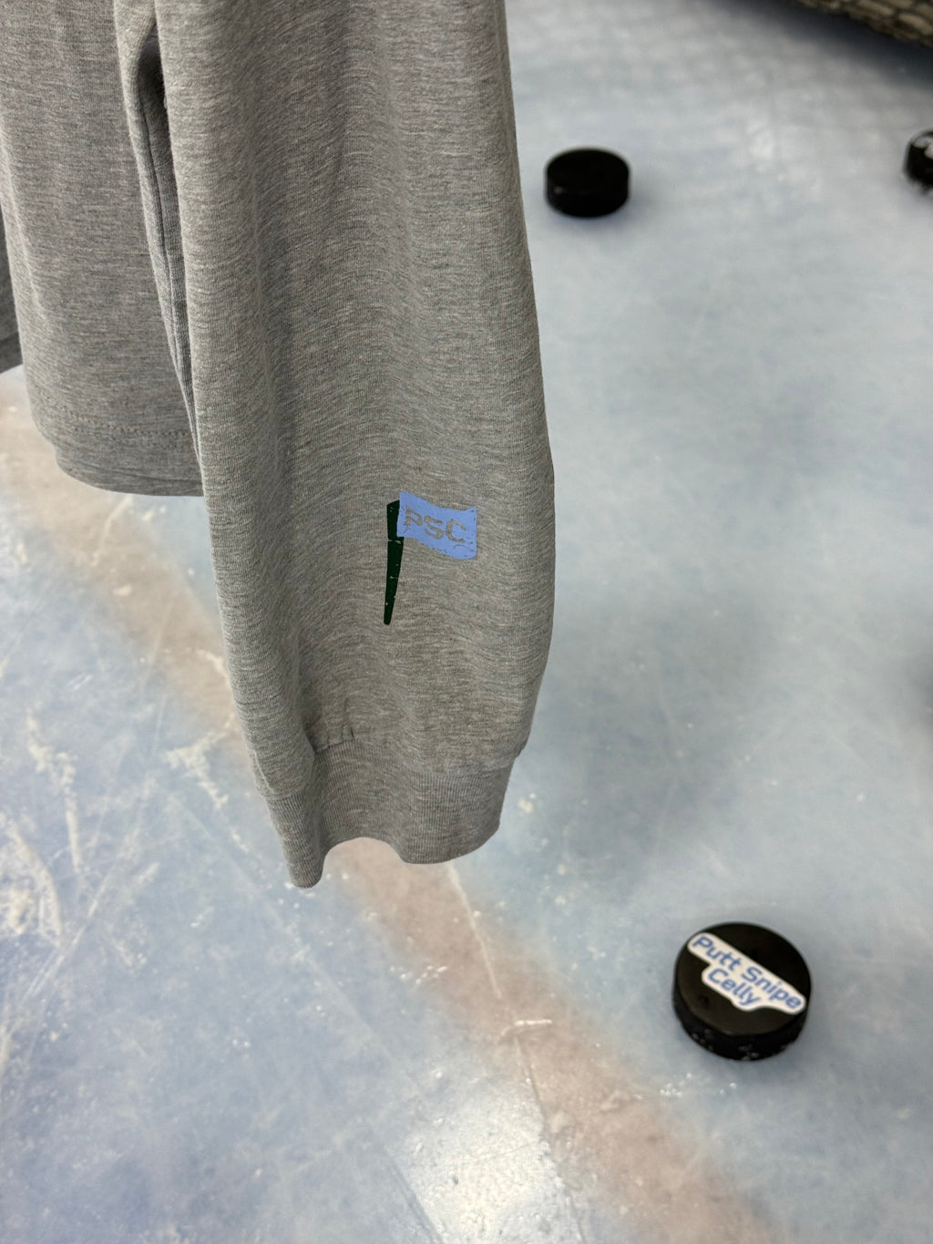 Close up sleeve featuring a view of the PSC icon logo on the athletic heather colored long sleeve lifestyle t-shirt shown hanging from a hockey net with pucks on the ice below. Presented by Putt Snipe Celly