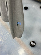 Close up sleeve featuring a view of the PSC icon logo on the athletic heather colored long sleeve lifestyle t-shirt shown hanging from a hockey net with pucks on the ice below. Presented by Putt Snipe Celly